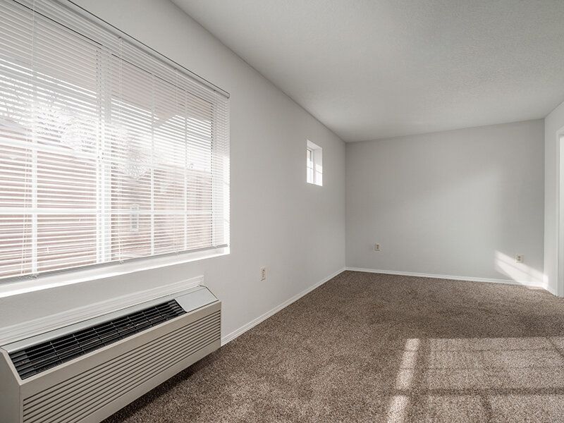 Empty apartment living room with beige carpet, large window with blinds, and wall-mounted heater at Santa Fe Suites in Santa Fe, NM.