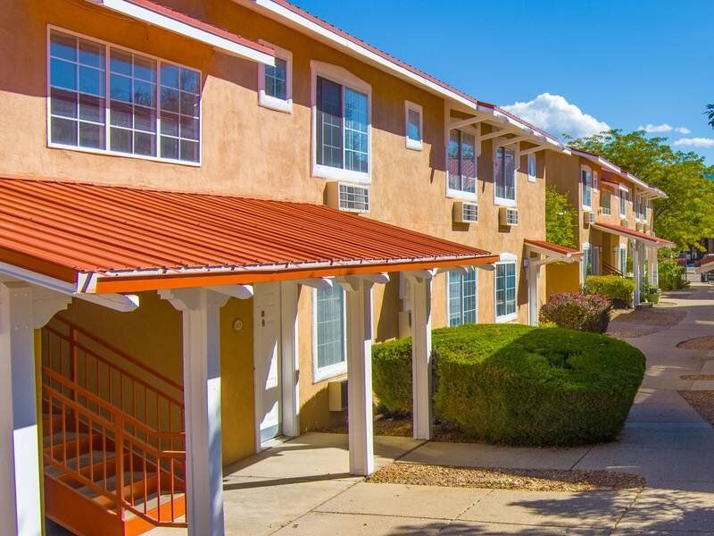 Exterior view of a beige apartment building with a red roof, white trim, and a walkway lined with shrubs at Santa Fe Suites in Santa Fe, NM.