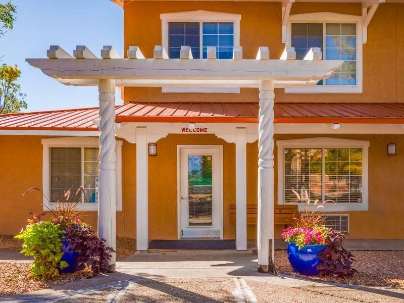 Exterior of apartment building with a covered entrance, white twisted columns, and potted plants at Santa Fe Suites in Santa Fe, NM.