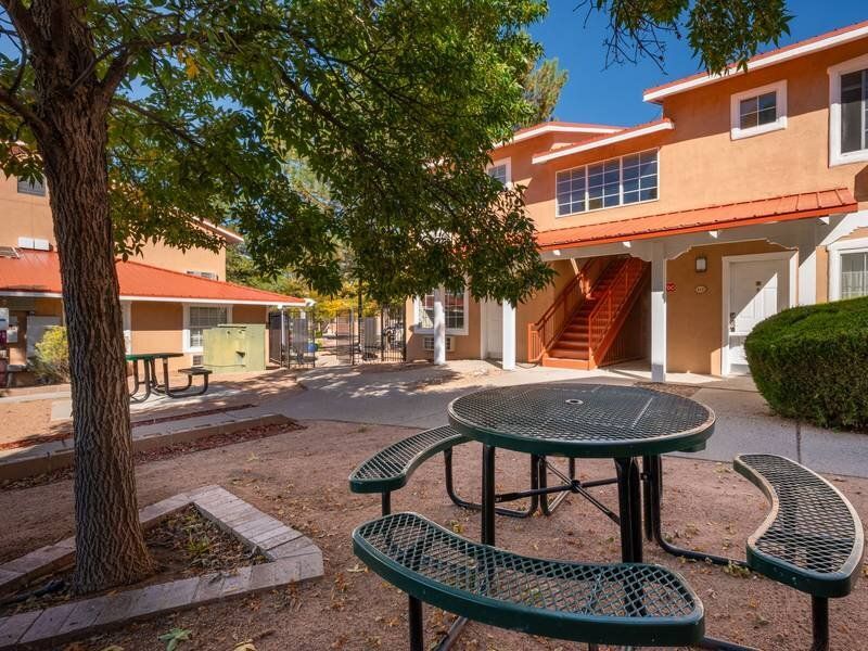 Communal courtyard at an apartment complex with orange buildings, a round metal picnic table, and trees at Santa Fe Suites in Santa Fe, NM.