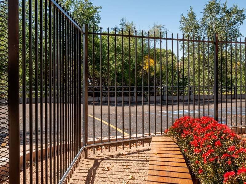 Tall black metal fence surrounding a paved parking area with red flowering bushes and trees at Santa Fe Suites in Santa Fe, NM.