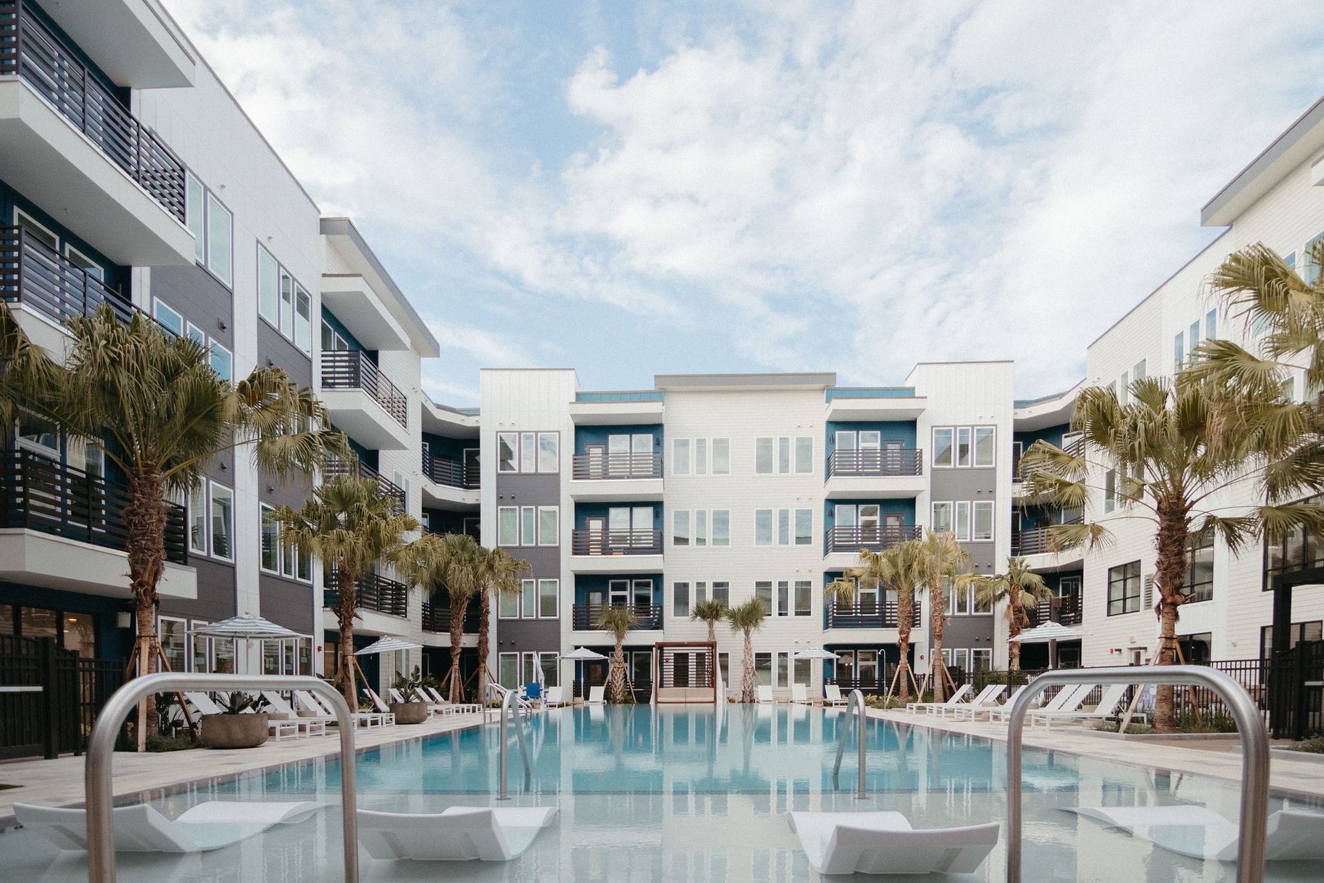 A courtyard with a swimming pool surrounded by modern apartment buildings and palm trees under a bright blue sky.