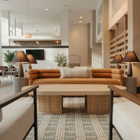 A modern, neutrally-toned lounge area with a tan sofa, wooden coffee table, and symmetrical lamps in a spacious lobby.
