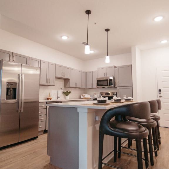 A modern kitchen with grey cabinetry, stainless steel appliances, a center island, and dark bar stools.