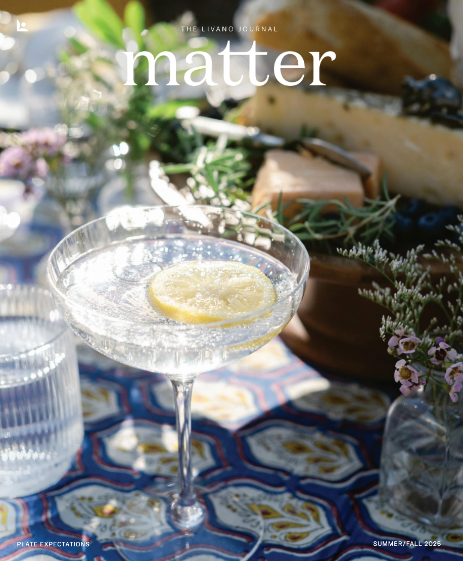 A cocktail glass with a lemon slice sits on a patterned blue tablecloth next to flowers and a cheese board.