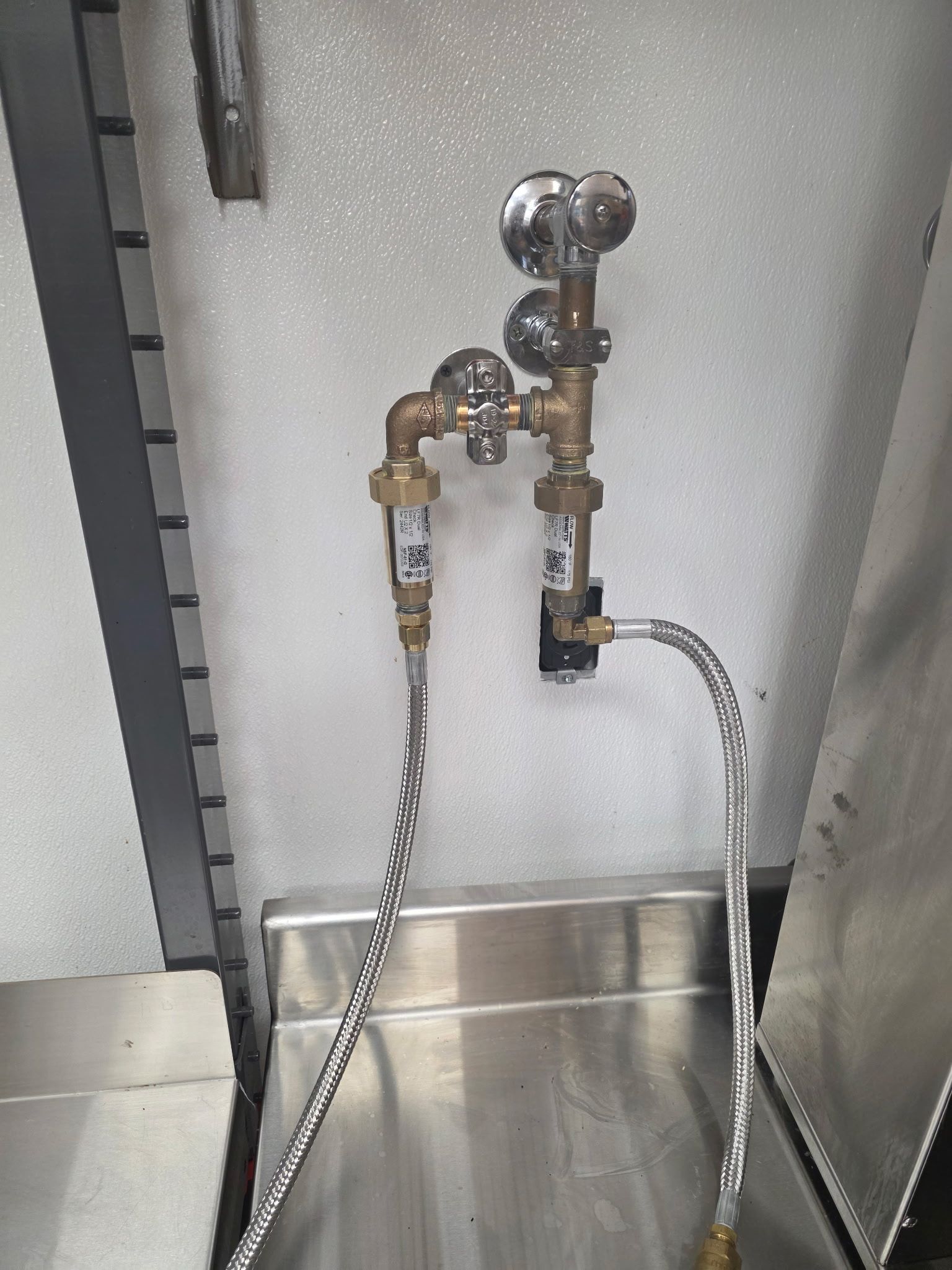 Plumbing fixtures, including valves and braided hoses, mounted above a stainless steel sink in a commercial kitchen.