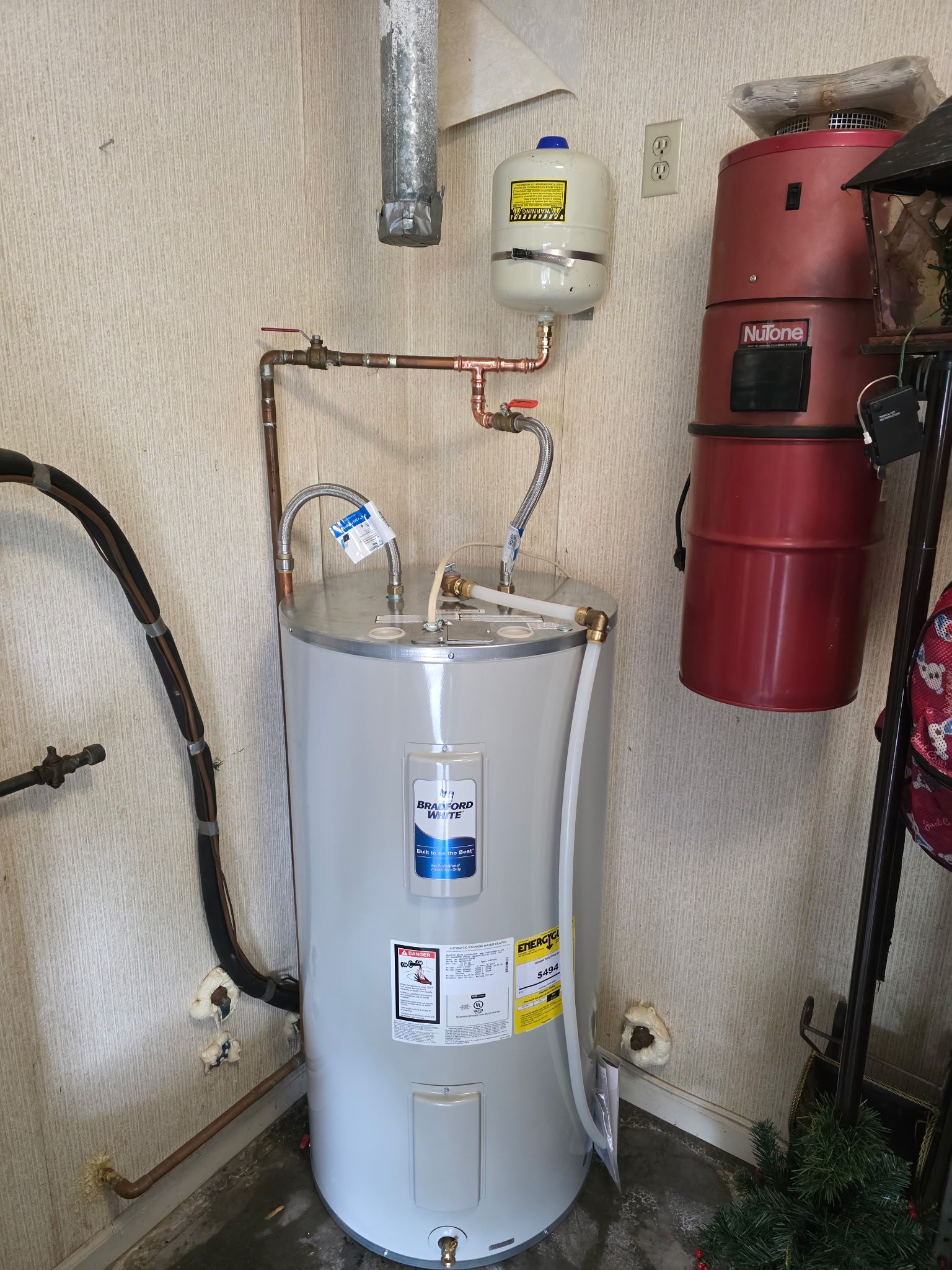 A water heater with copper pipes, a pressure tank, and a central vacuum unit in a utility room.