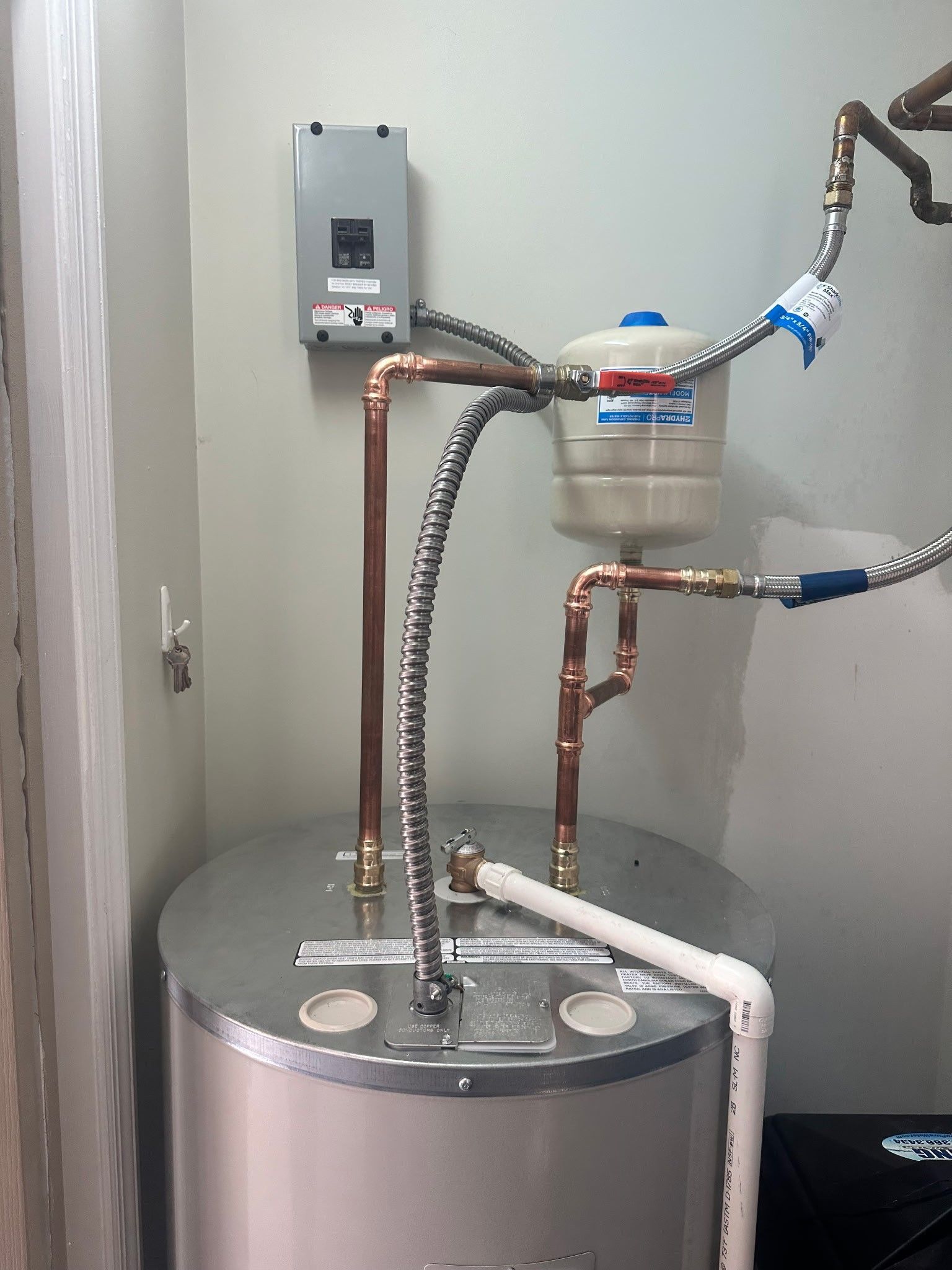 Water heater with copper pipes, a pressure tank, and electrical box, against a light green wall.
