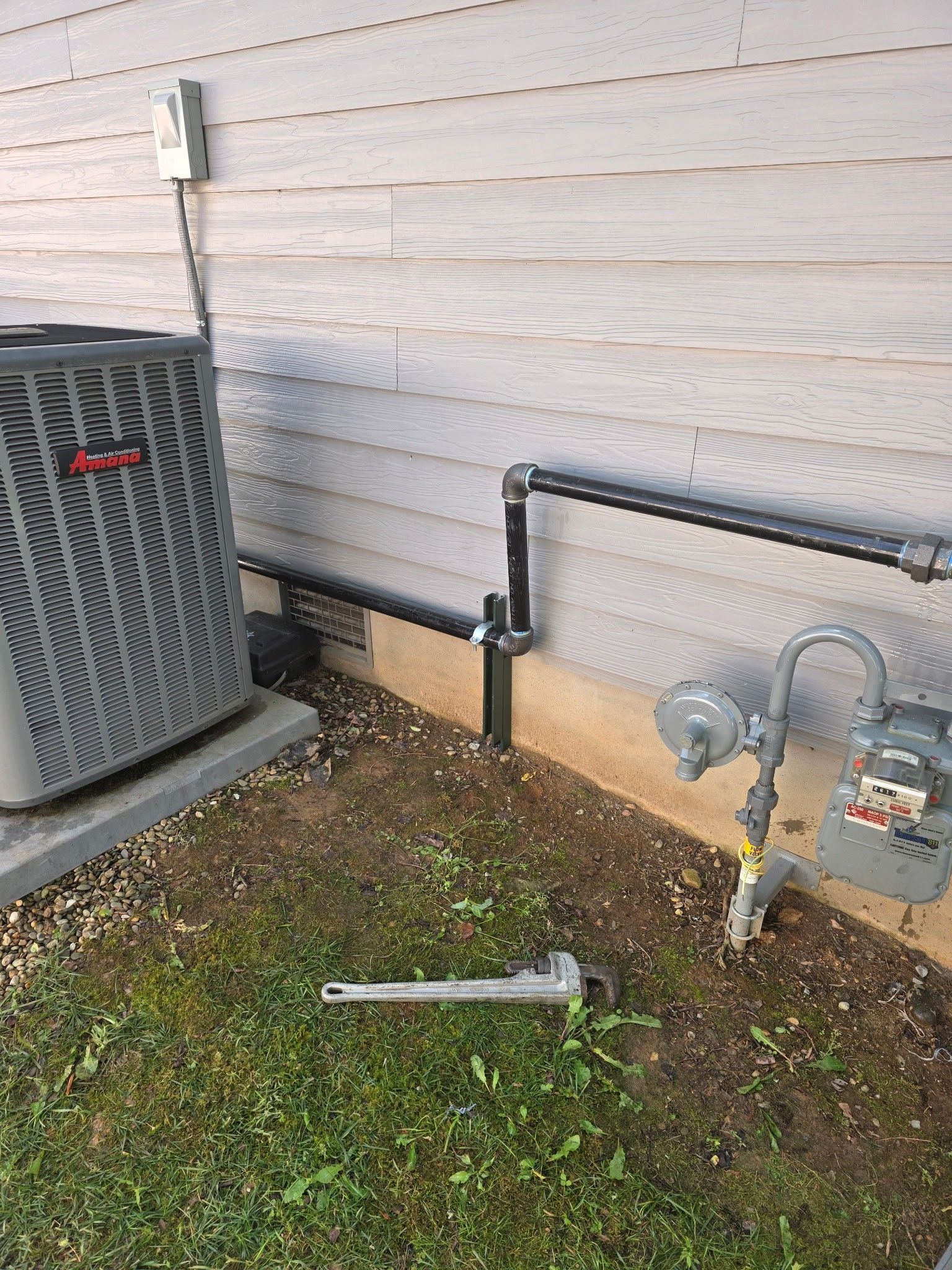AC unit and gas meter outside a building with exposed pipes, with a wrench resting on the ground.
