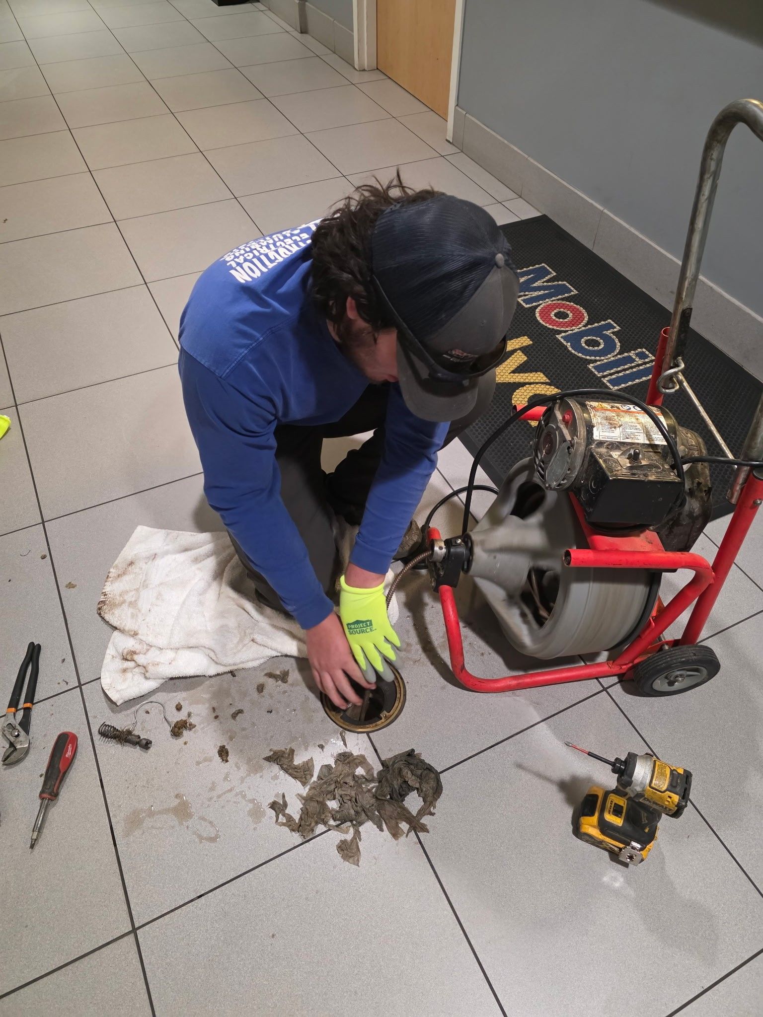 Plumber clearing a drain. Indoors. Person in blue shirt, black cap, and gloves, working near a drain.