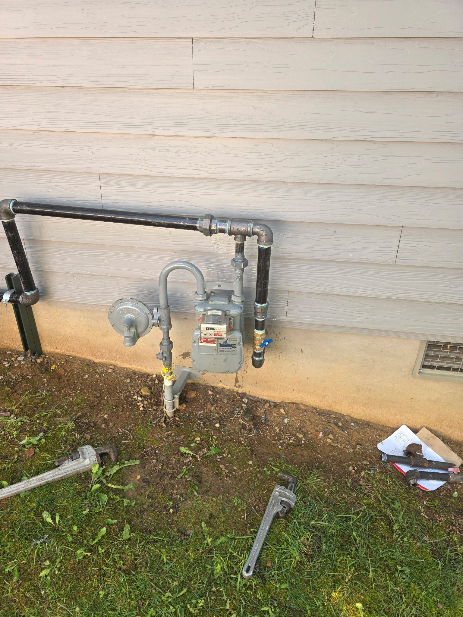Gas meter on a building's exterior with black pipes, wrenches on the ground, and beige siding.