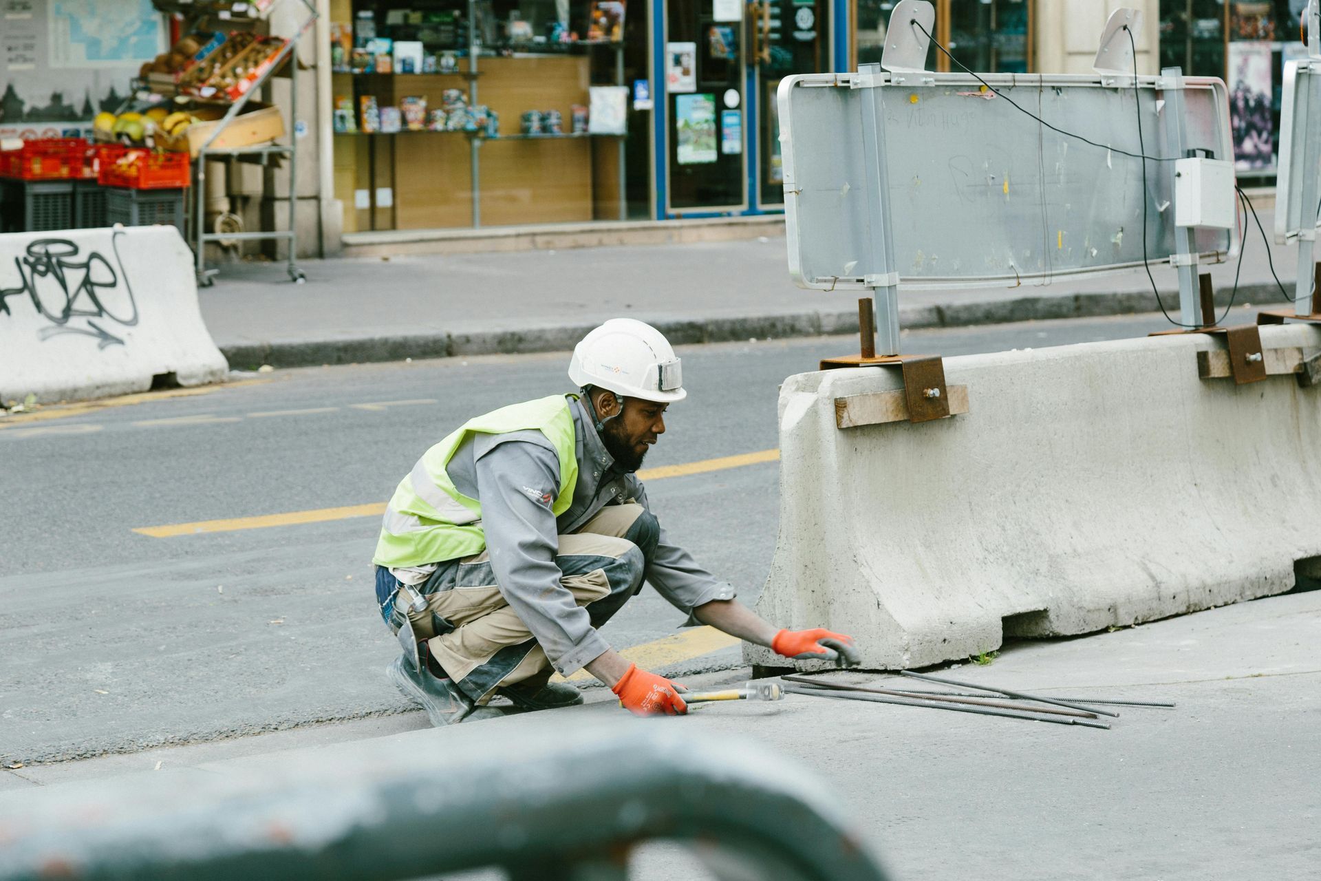 Construction worker kneels on a street, orange gloves, white hard hat, high-visibility vest, concrete barriers.