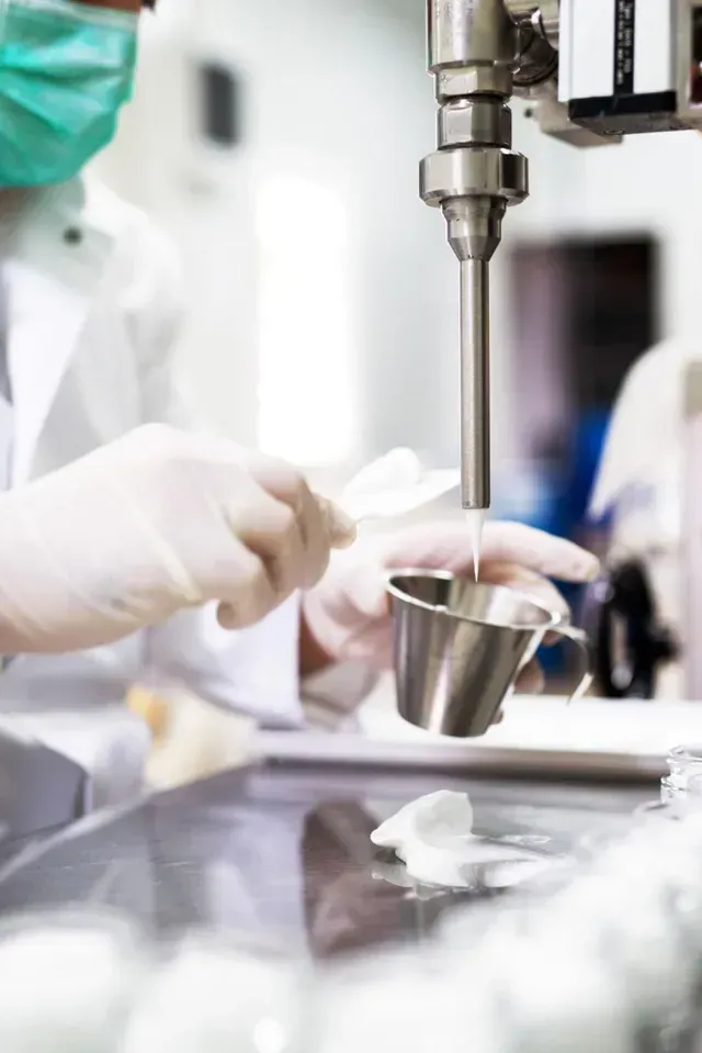 Gloved worker pouring liquid from a metal cup under a machine in a laboratory