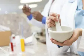 Person grinding ingredients in a mortar and pestle in a lab, with test tubes in the background