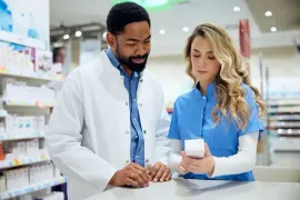 Two pharmacy staff review a prescription at a counter, one in a white coat and one holding a medication bottle.