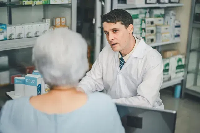 Pharmacist speaking with a customer at a pharmacy counter, shelves of medicine in the background.