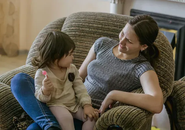 Woman and child sitting together in a cushioned chair, smiling at each other in a cozy room