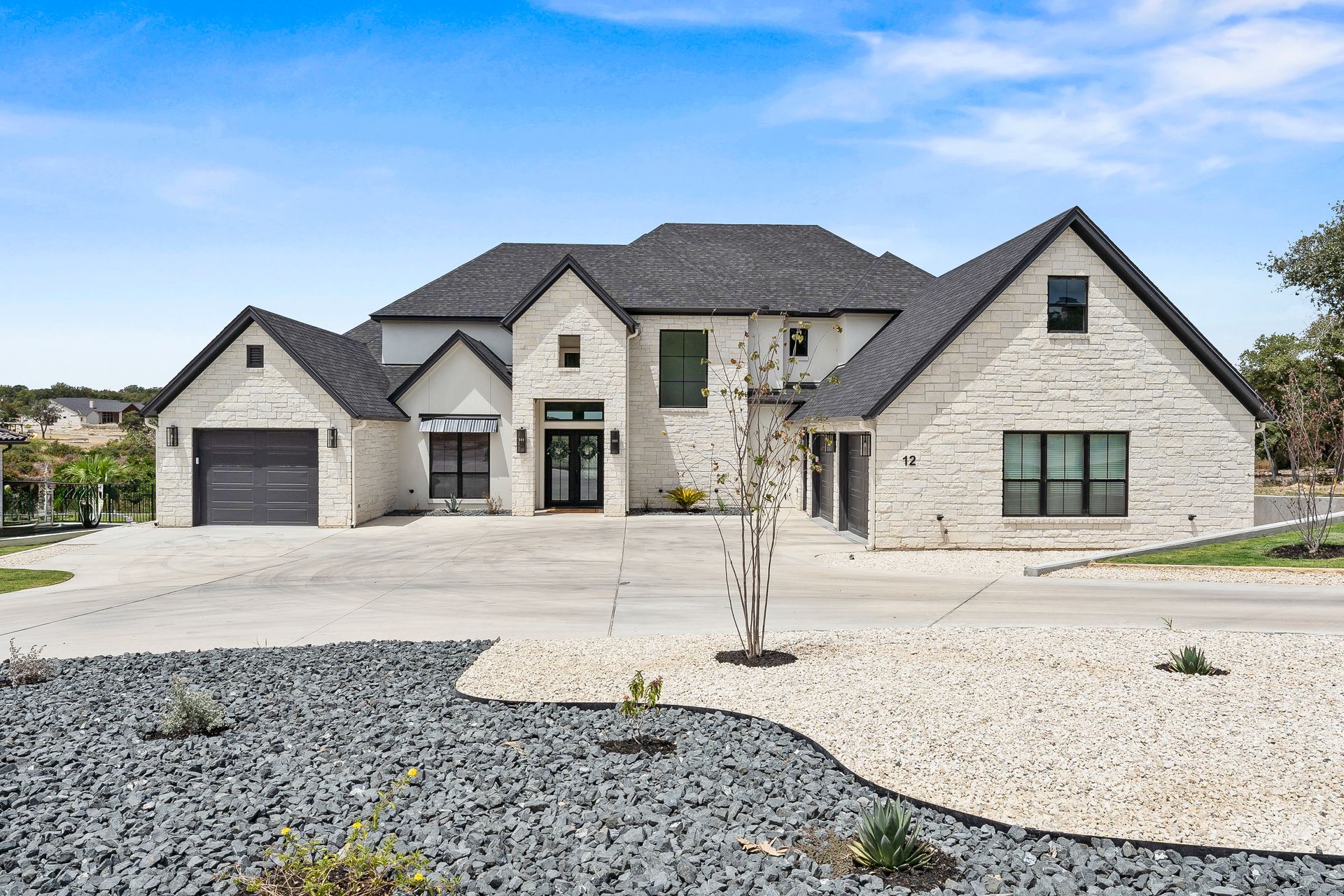 Modern stone-and-brick suburban house with a dark roof, garage, and gravel front yard under a blue sky