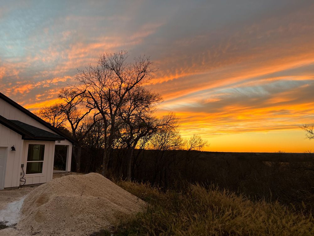 Sunset over a rural yard with a house, bare tree silhouettes, and a mound of gravel under orange clouds