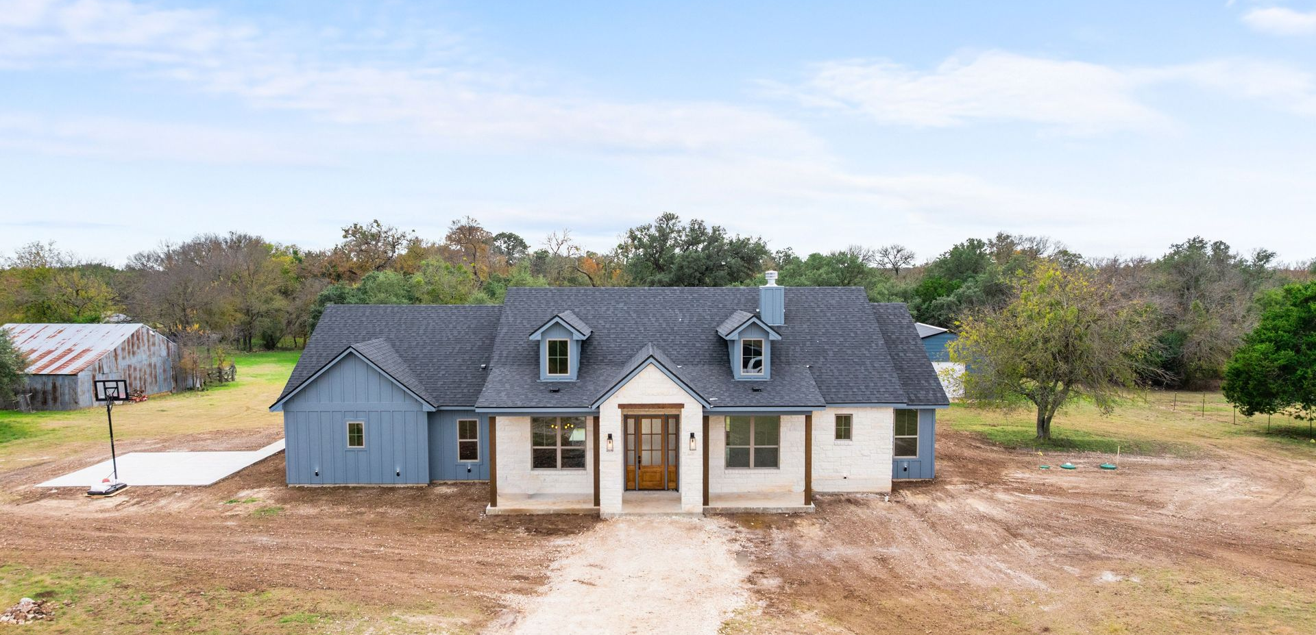 Single-story gray house with a dark roof and front porch on a bare lot with trees behind it
