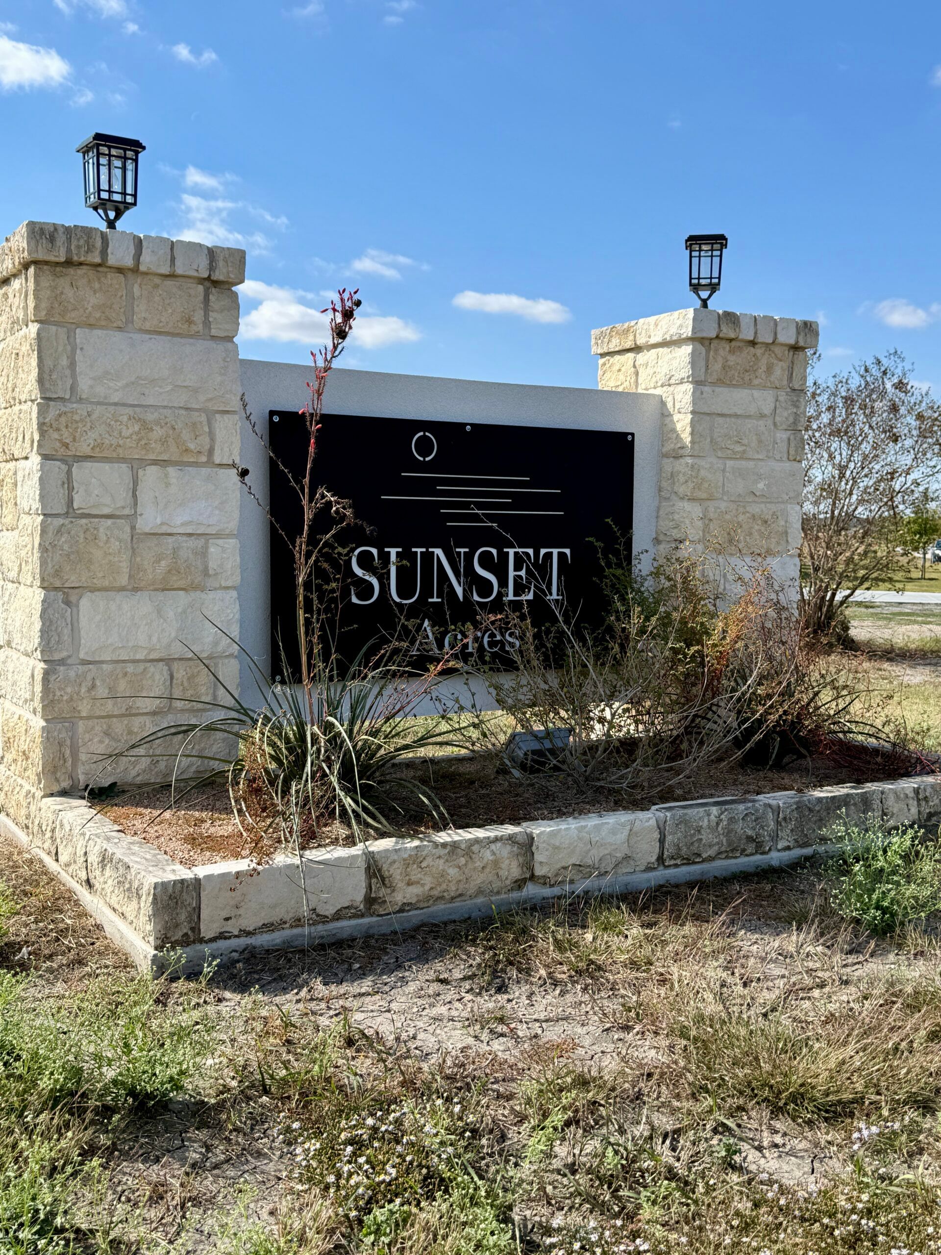 Stone entrance sign for Sunset community; black sign with white text, lamps on top, blue sky background.