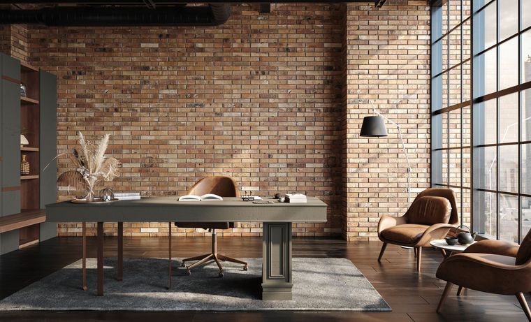 A modern home office with a gray desk on a rug, brick walls, two brown armchairs, and floor-to-ceiling industrial windows.