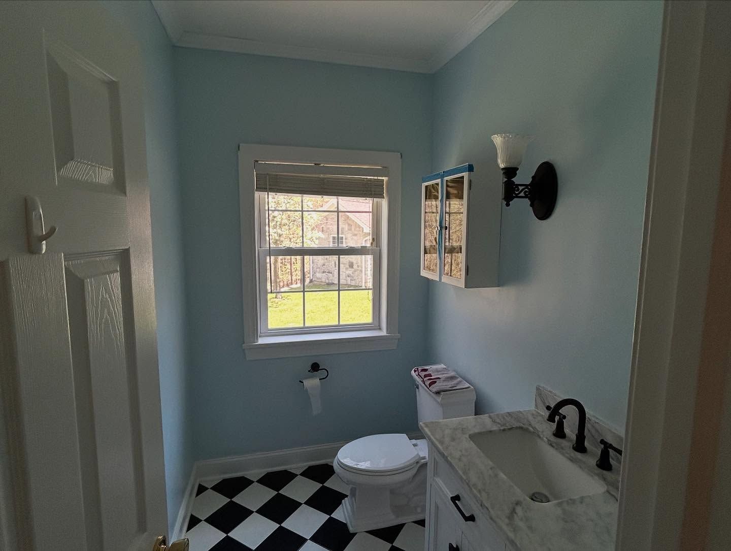 Blue bathroom with checkered floor, toilet, sink, and window. Black sconce and cabinet.