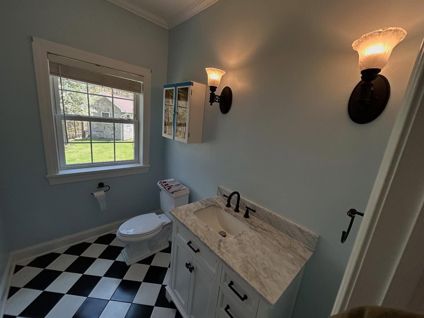 Bathroom with black and white checkered floor, blue walls, a window, toilet, vanity, and wall sconces.