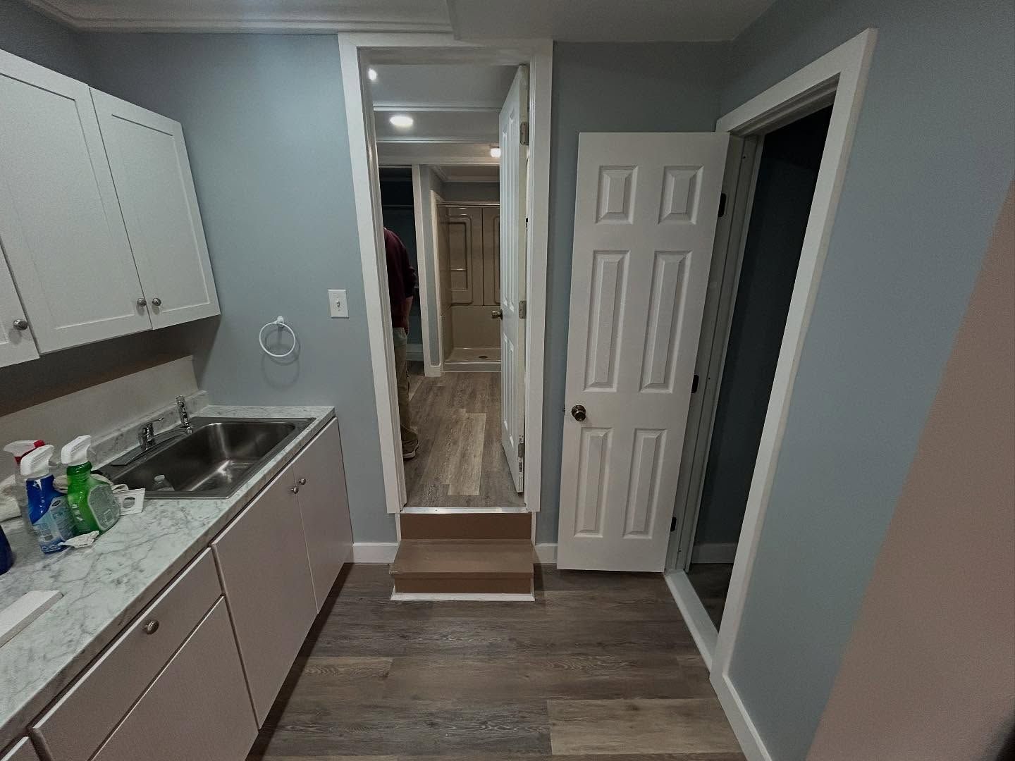 A laundry room with gray walls, white cabinets, and a sink. A doorway leads to a hallway with a wood floor.