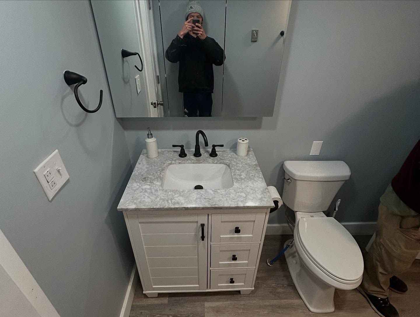 Bathroom with a gray vanity, white countertop, and a white toilet. A person is visible in the mirror.
