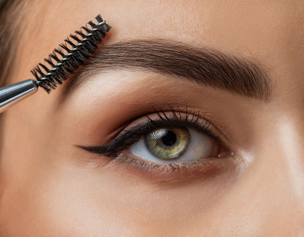 Primo piano di un occhio con uno scovolino per mascara che applica mascara scuro su ciglia e sopracciglia.