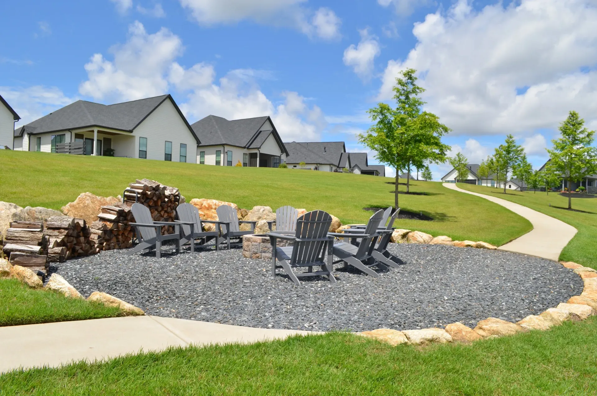 Stone fire pit with chairs in a landscaped suburban community park, beside a winding path and houses.