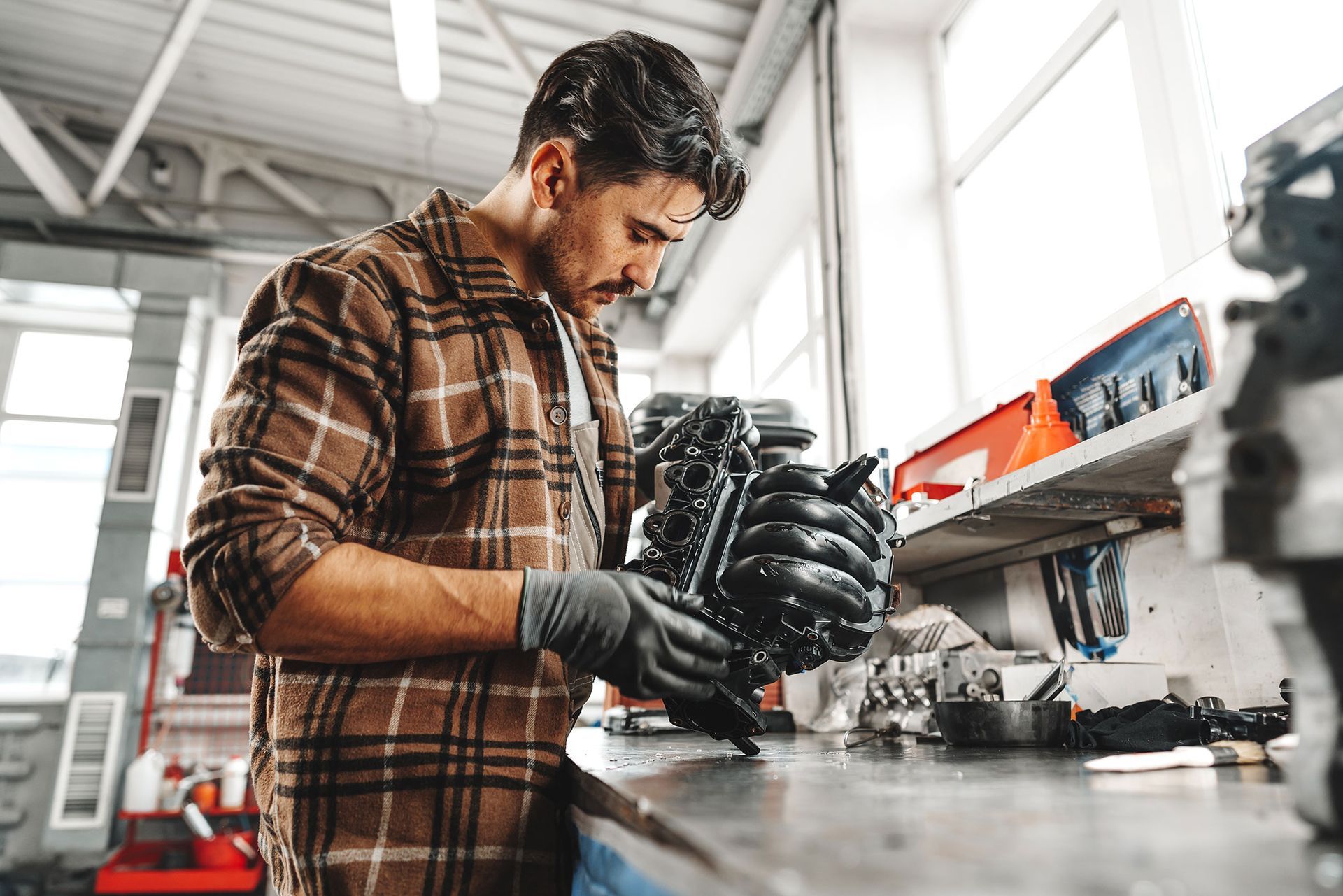 A mechanic wearing a brown plaid shirt and gloves works on a car engine component at a workbench in a workshop.