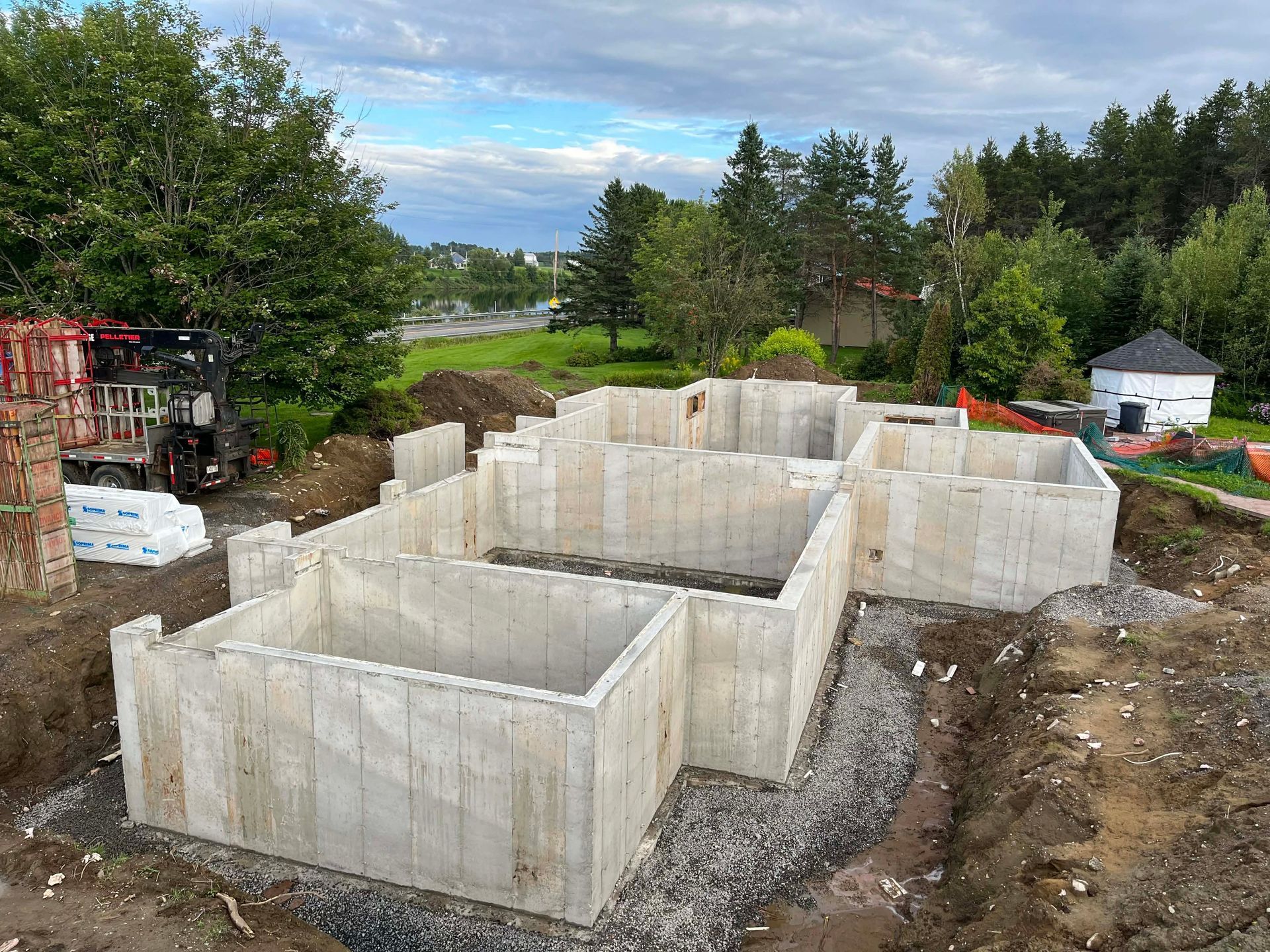 Murs de fondation en béton en construction sur un terrain dégagé, avec des arbres et des maisons en arrière-plan.
