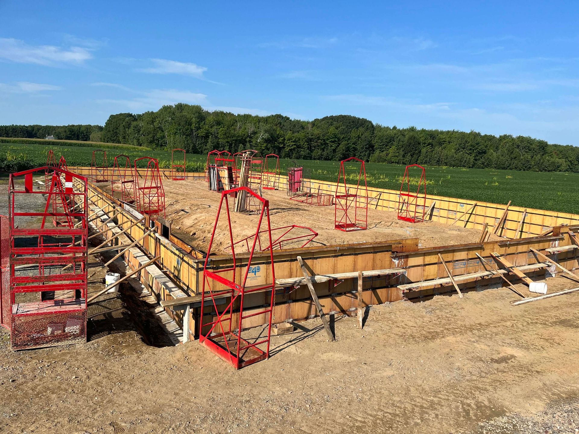 Chantier de construction avec coffrage de fondation en bois et échafaudages de sécurité rouges sous un ciel bleu clair