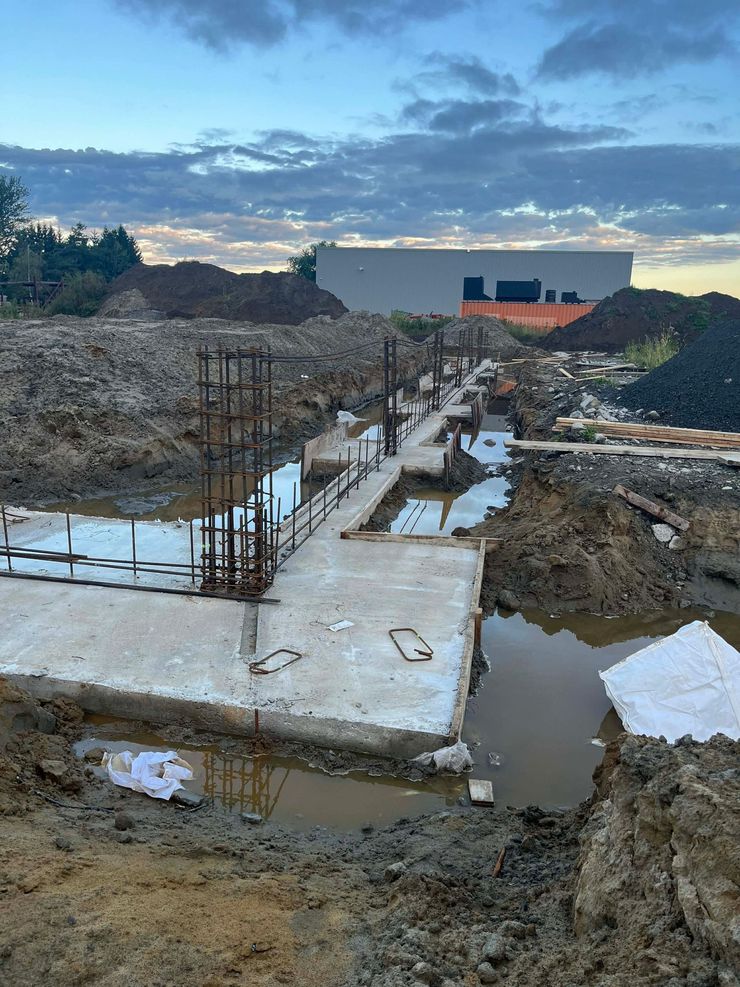 Fondations en béton en construction sur un terrain boueux, avec des barres d'armature, des ouvriers et un bâtiment en arrière-plan.