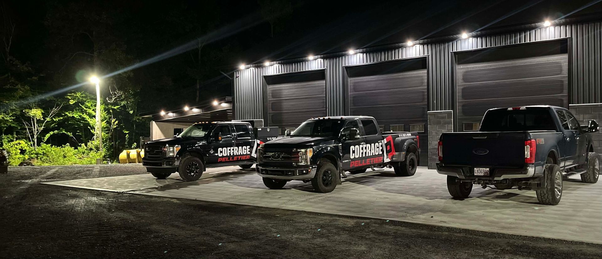 Trois camions tout-terrain stationnés de nuit devant un garage industriel éclairé.