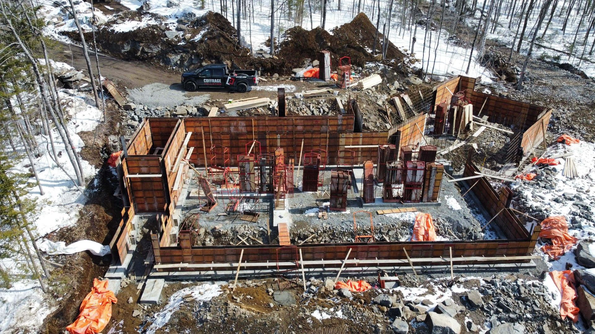 Vue aérienne d'un chantier de construction enneigé avec une structure en acier couleur rouille et des excavatrices.
