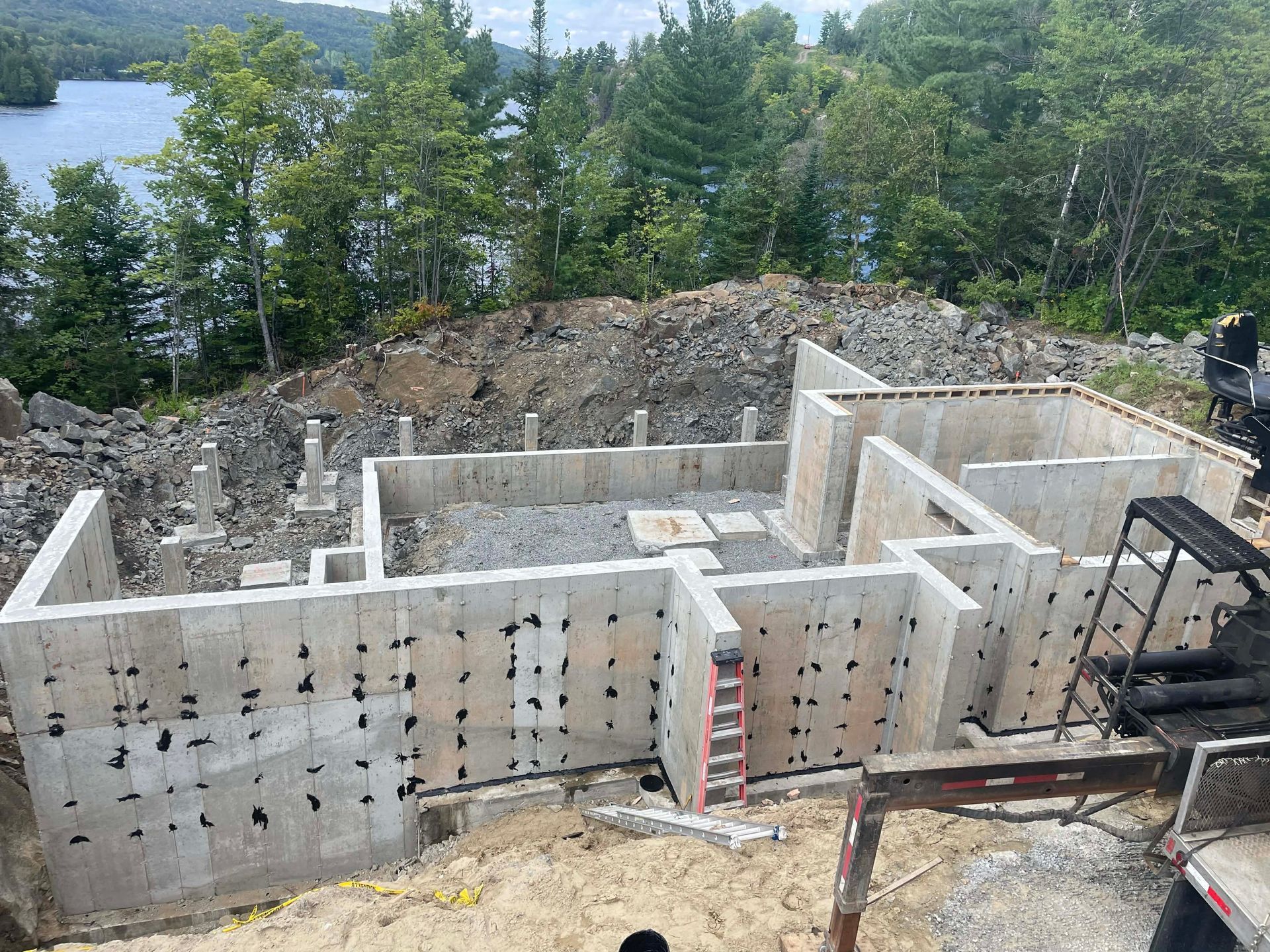 Des murs de fondation en béton sont en construction sur un flanc de colline boisé, avec une échelle et des engins de chantier à proximité.