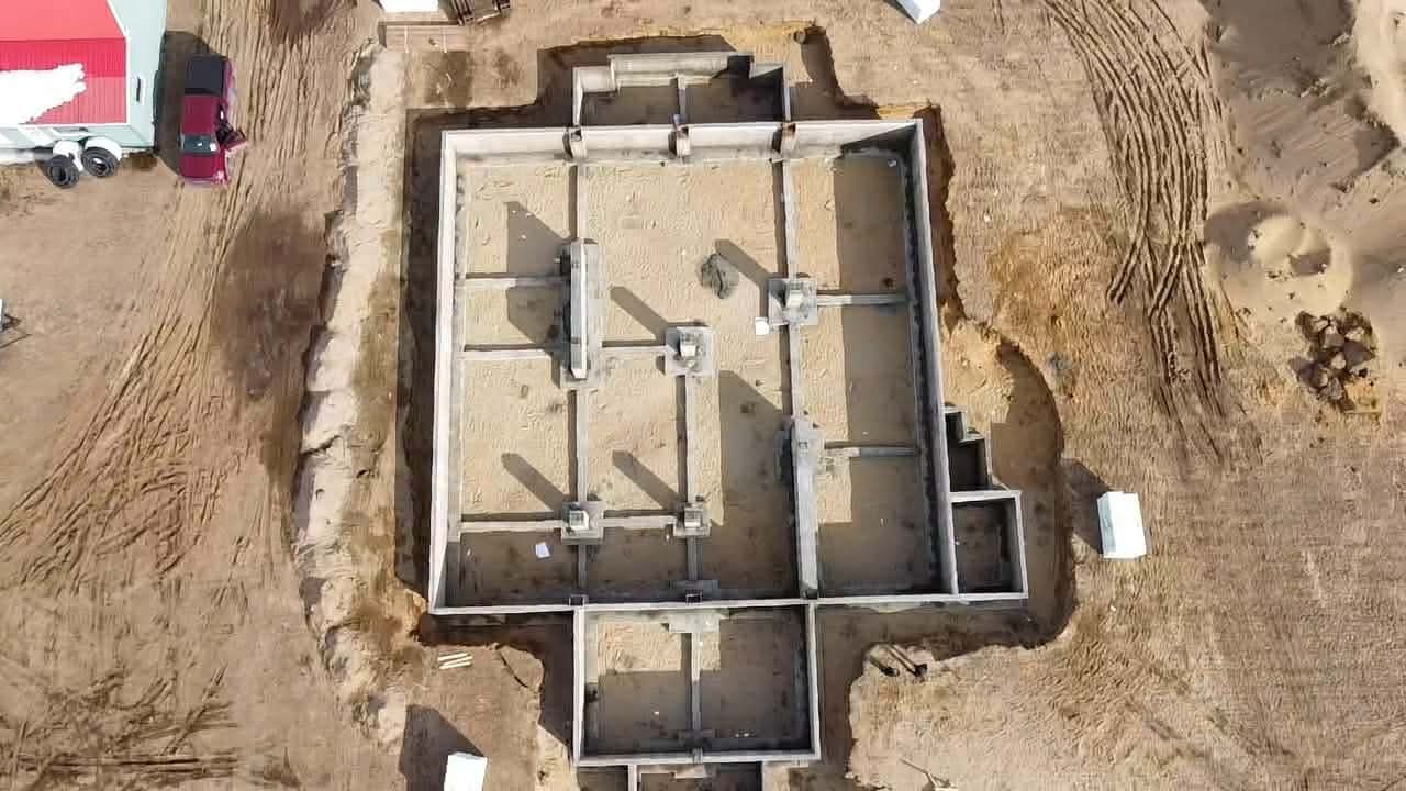 Vue aérienne des fondations d'une maison en béton sans toit, sur un terrain en terre battue, avec des équipements et des véhicules à proximité.