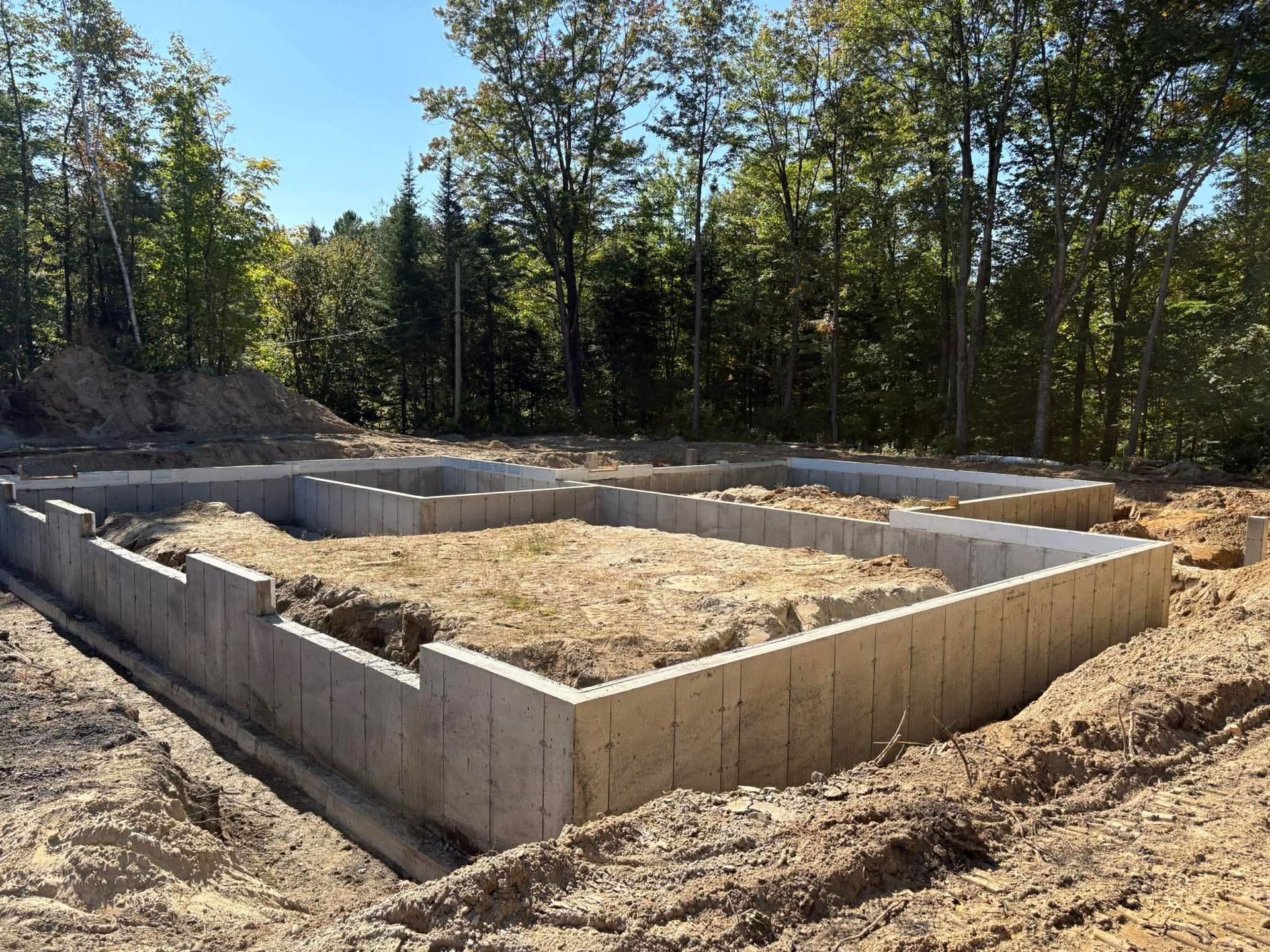 Des murs de fondation en béton sont en construction dans une clairière boisée, entourés de terre et d'arbres.