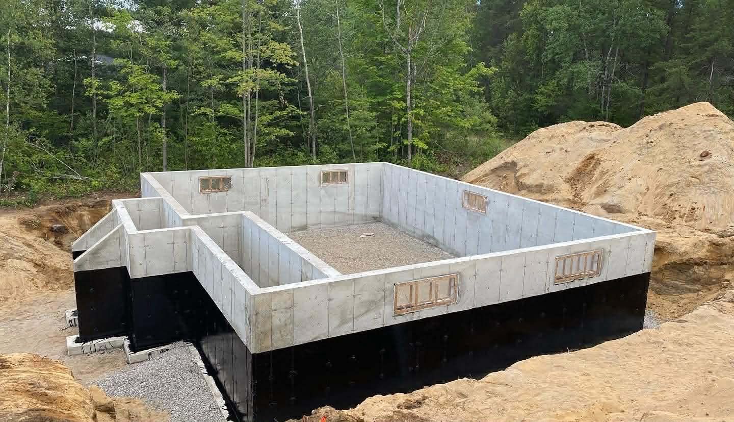 Murs de fondation en béton en construction sur un terrain boisé, avec des tas de terre et une membrane d'étanchéité noire à l'extérieur.