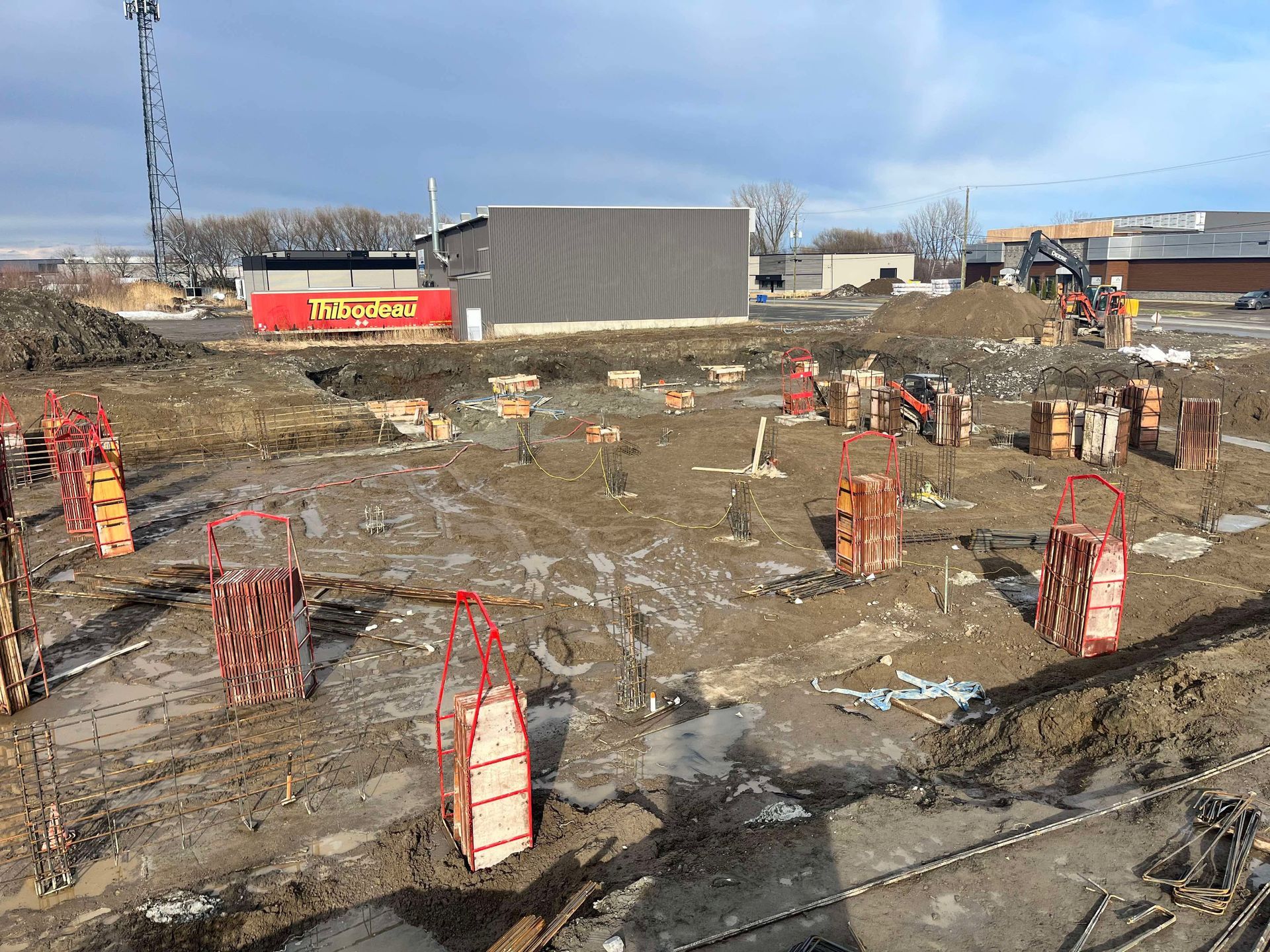 Chantier de construction avec coffrages de fondation en béton et armatures sur un sol boueux sous un ciel radieux