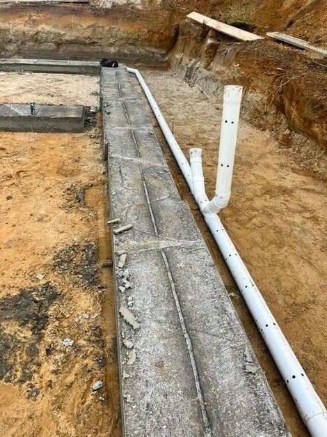 Tranchée avec semelle en béton et tuyaux en PVC blanc dans une fondation en terre excavée.