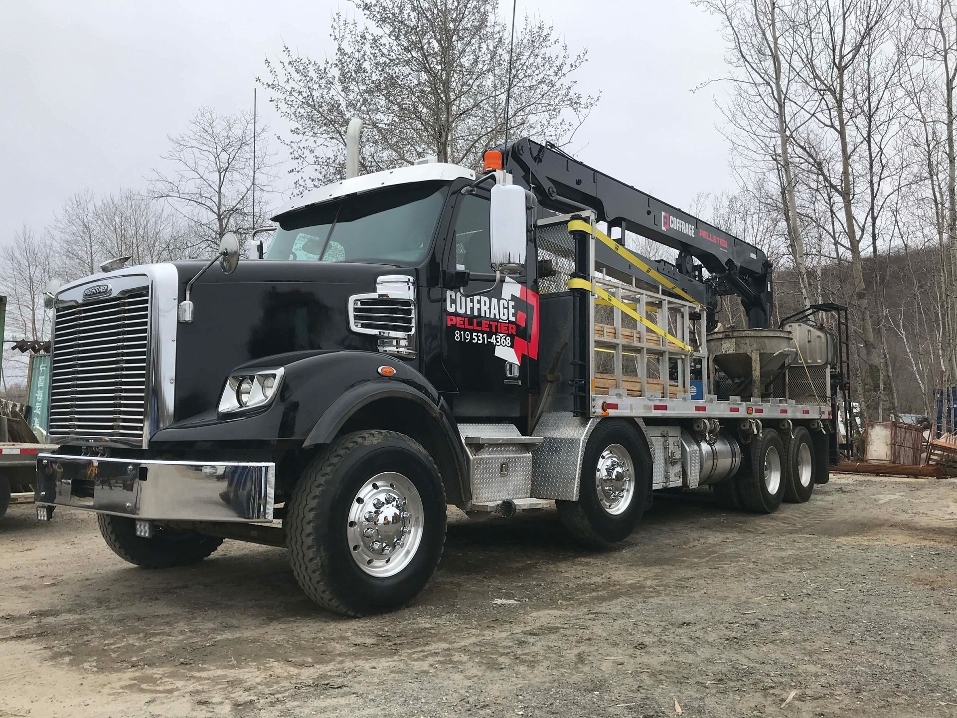 Camion-grue commercial noir stationné sur du gravier, flèche déployée, arbres dénudés en arrière-plan