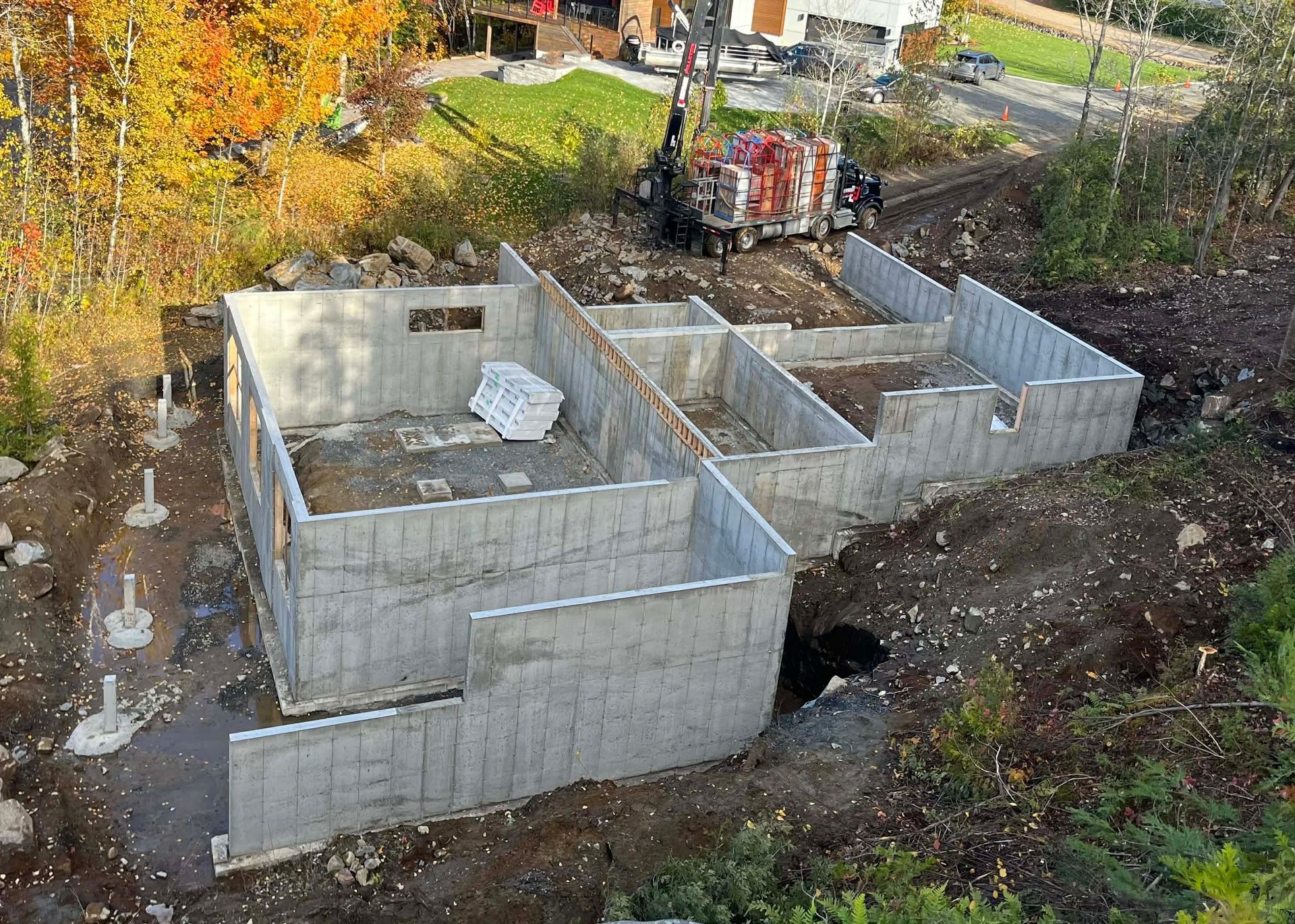 Des murs de fondation en béton sont en construction à flanc de colline, avec des arbres d'automne et un camion à proximité.