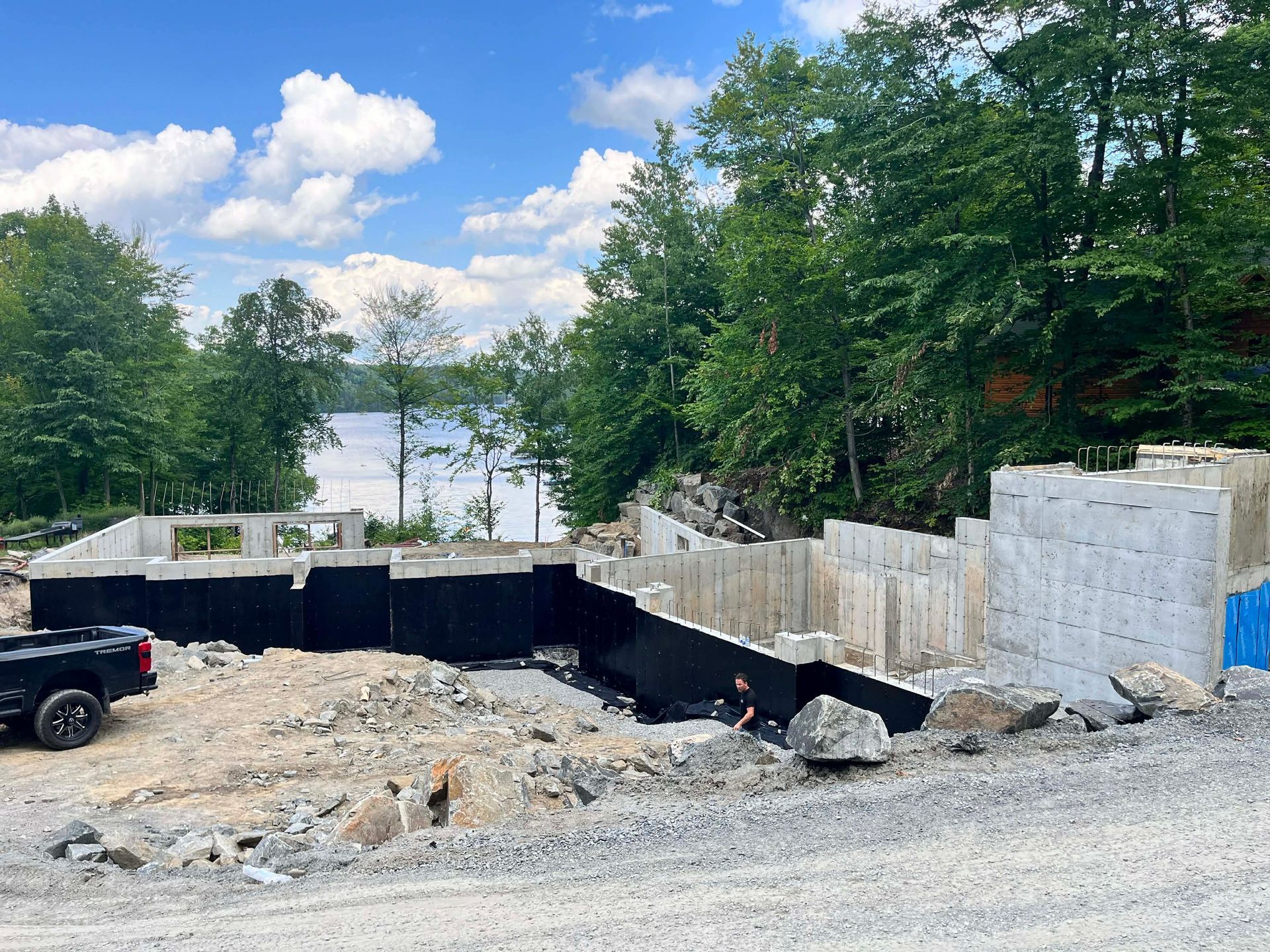 Chantier de construction avec fondations excavées, mur de soutènement noir, gravier et arbres au bord d'un lac