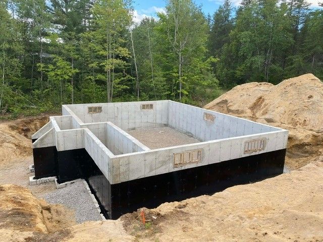 Murs de fondation en béton sur un chantier de construction boisé, avec des tas de terre autour de la fouille.