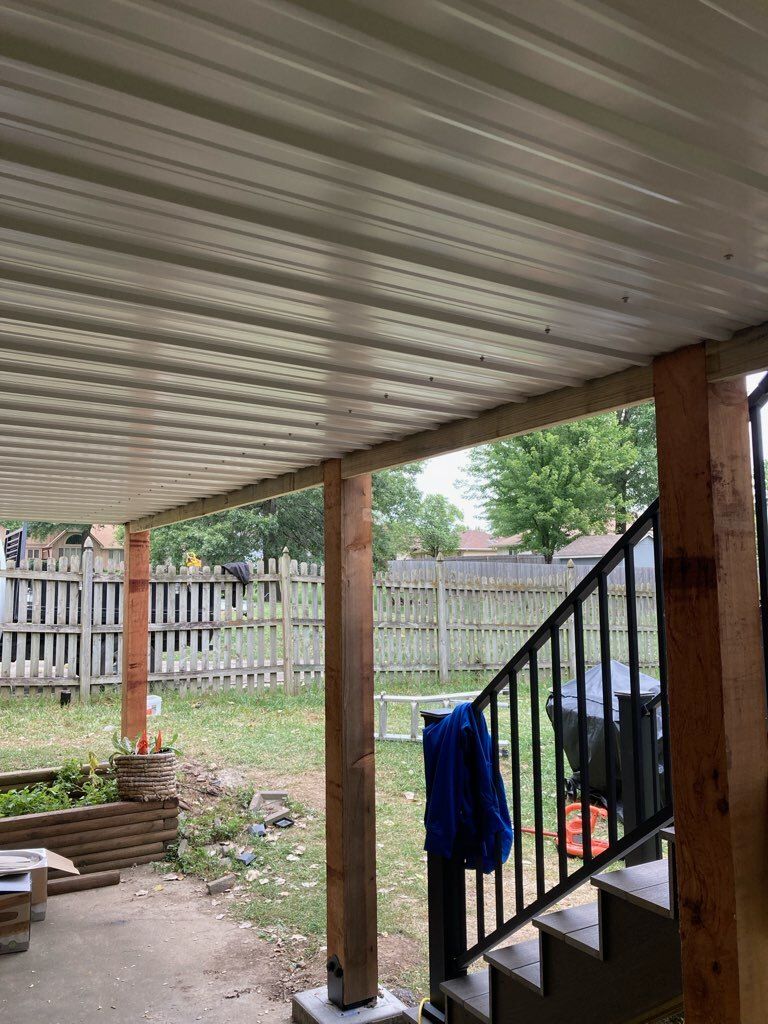 Covered patio with metal roof supported by wood posts, overlooking a grassy backyard with a fence.