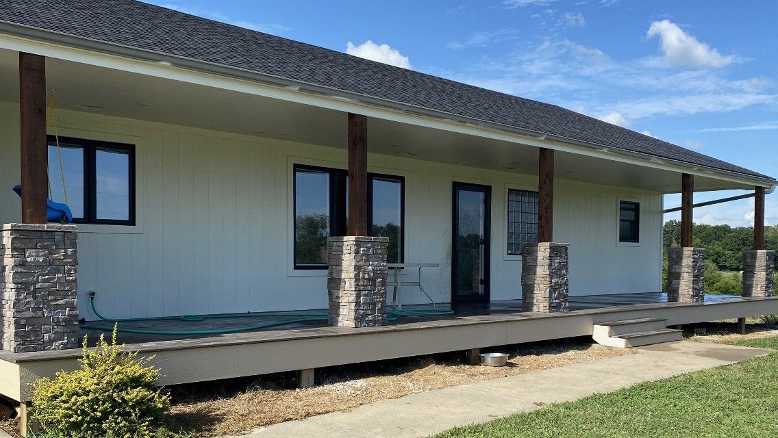 Tan house with dark roof and trim, wooden porch with stone columns, and green yard.